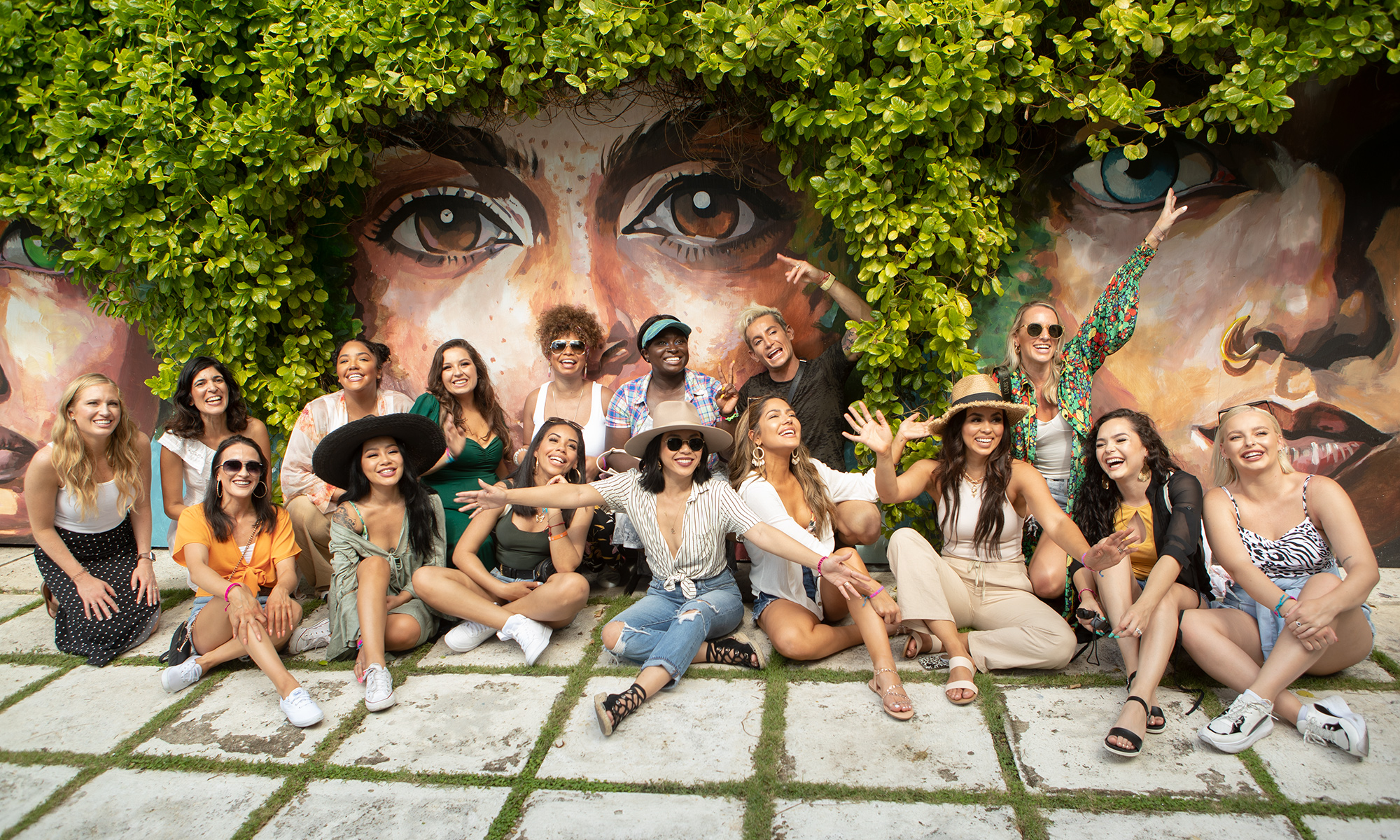 A diverse group of people are sitting outside on a well landscaped sidewalk in front of a mural of diverse faces in the Bahamas. They are all smiling as they take part in the award-winning Beautyscape influencer program.
