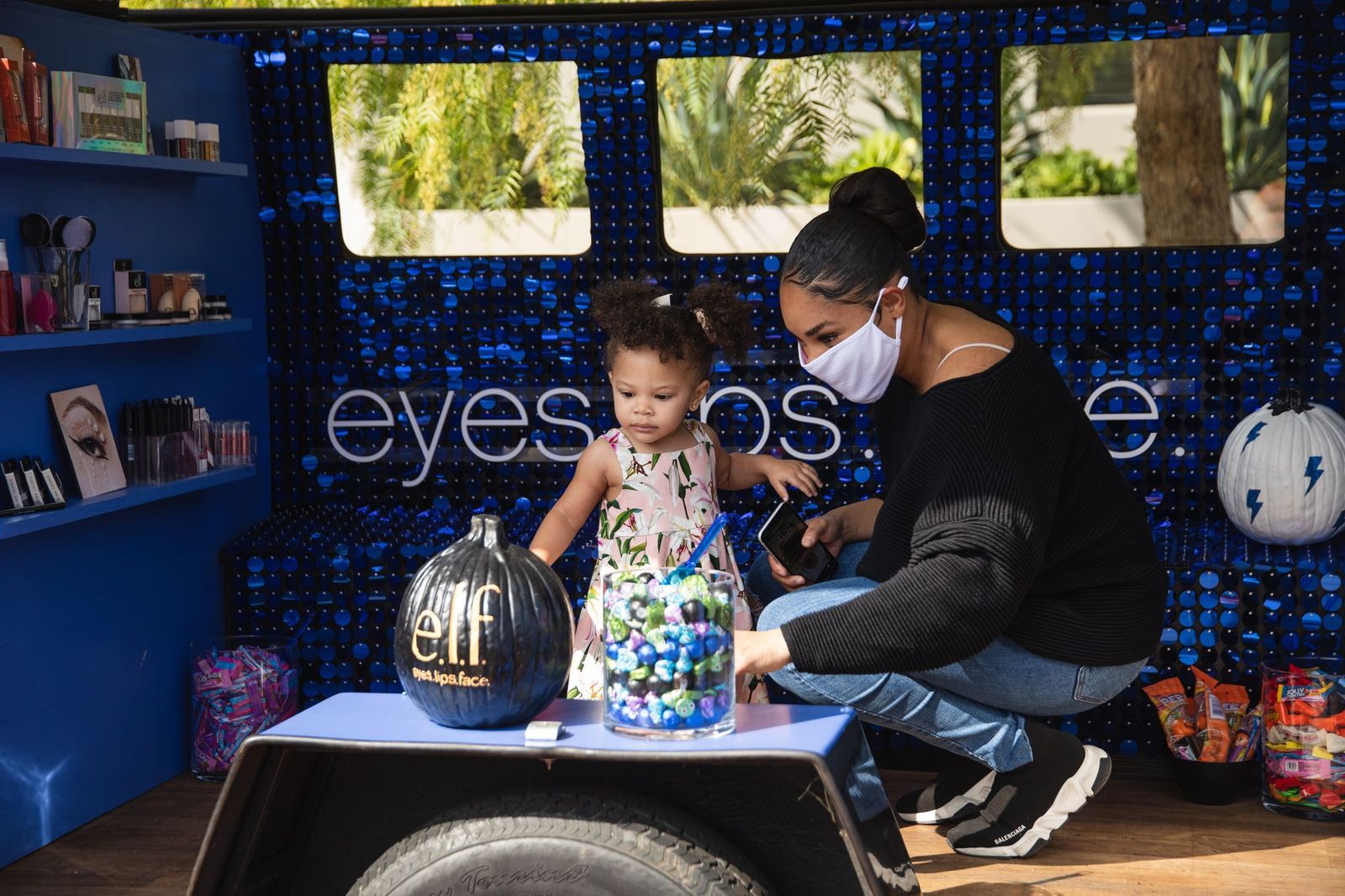 female presenting person wearing white mask with young female presenting child, sitting inside pop-up vehicle with purple sparkly wall behind her with the words "eyes lips face" in white.