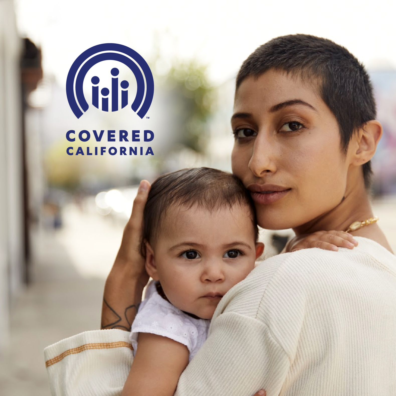 Female identified person with short-cropped brown hair, holding a baby to her chest. Covered California Logo overlays image in upper left corner.