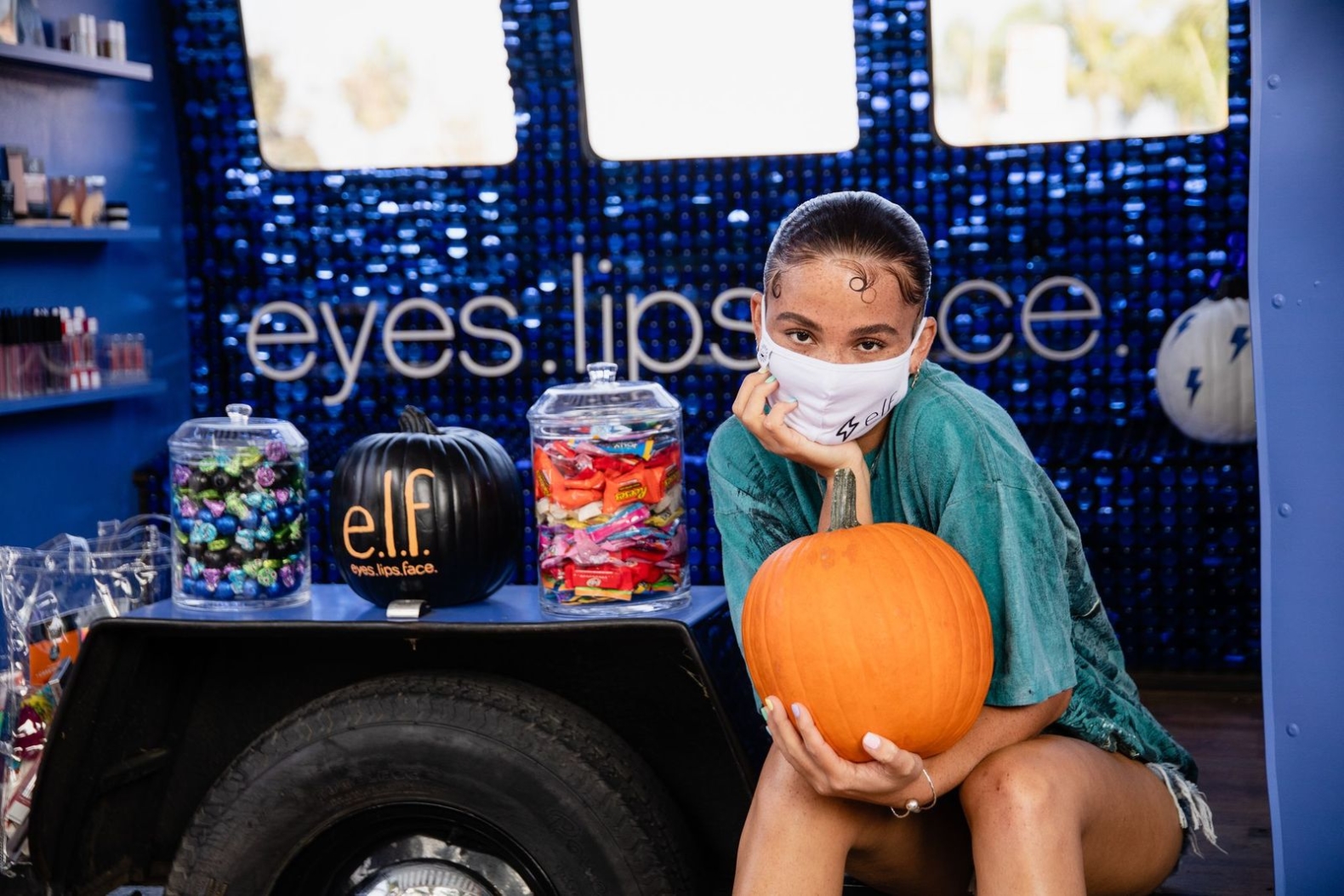 female presenting person wearing teal shirt and white e.l.f. branded face mask, holding orange pumpkin while sitting inside pop-up vehicle with purple sparkly wall behind her with the words "eyes lips face" in white.