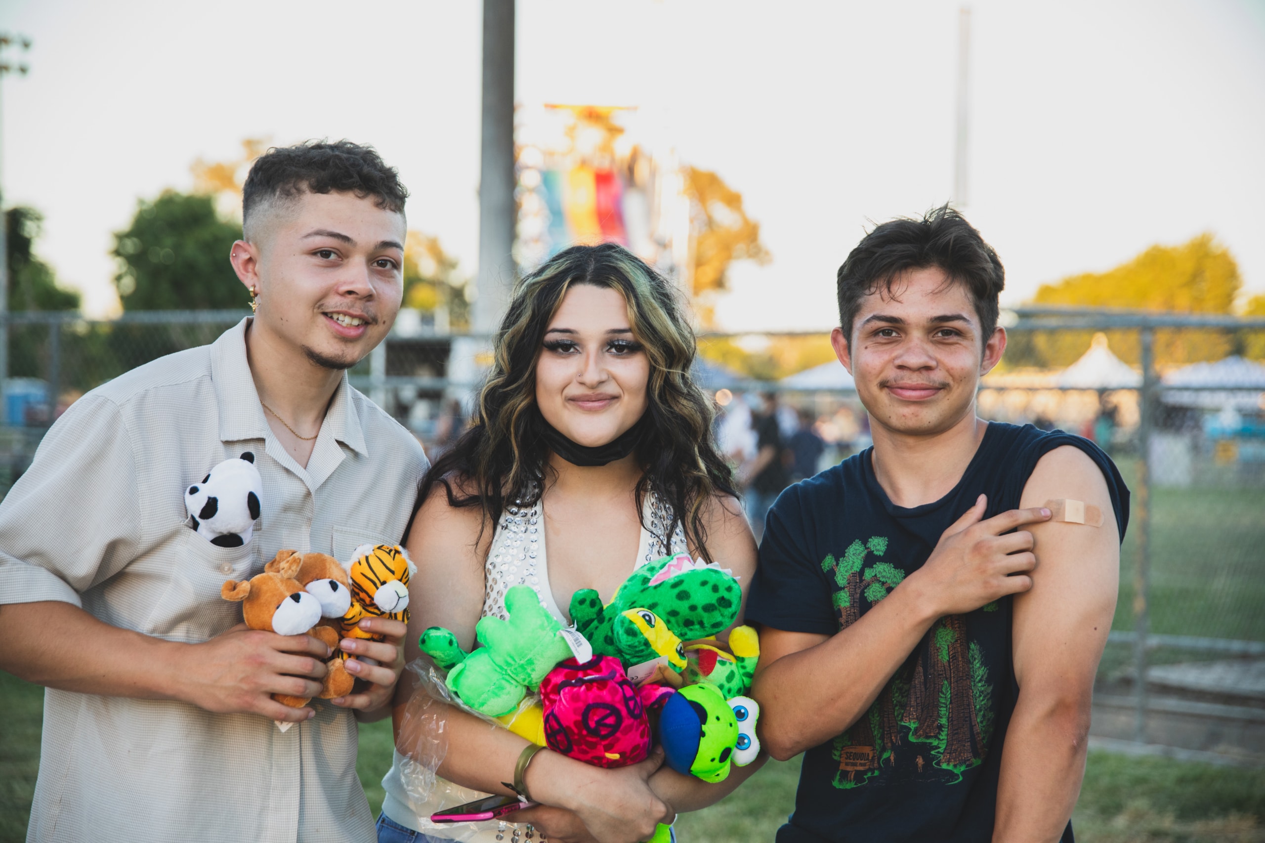Three people stand together smiling at a carnival. One proudly shows the bandaid on their recently vaccinated arm and the other two hold colorful prizes they won at the carnival. Two hold colorful stuffed animal prizes while the other lifts their shirt sleeve to show a bandage from a recently administered vaccine shot.