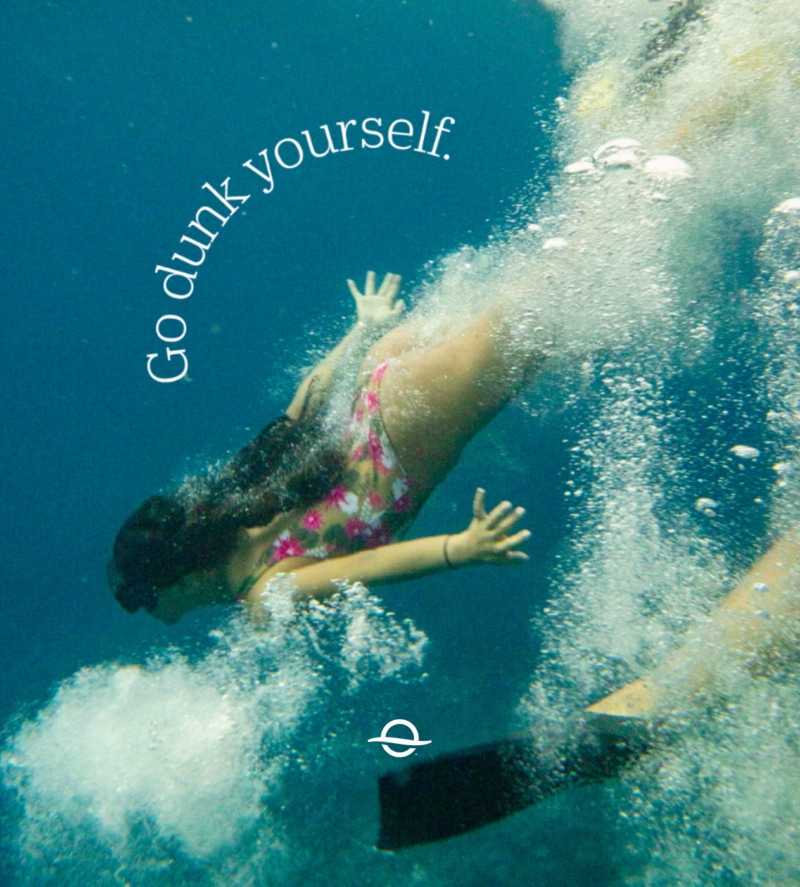 Photo image of a young girl diving into the ocean with bubbles framing the bottom right and side of the image. There is another person’s leg with a swimming fin coming out from the bottom right side. A curved headline near the top left in white type reads, “Go dunk yourself.” and the white Outrigger O Icon is at the center bottom.