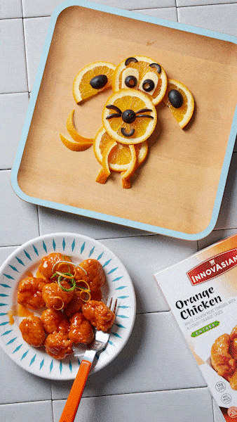 An animated gif of a small plate of orange chicken sit in the lower half of the frame, next to a box of InnovAsian brand Orange Chicken. Above it, there is a square wooden plate with an adorable, smiling food-art dog made out of orange slides and black olives. A small bone appears on the plate and the dog's eyes dart over to look at it excitedly while their tail begins to wag.