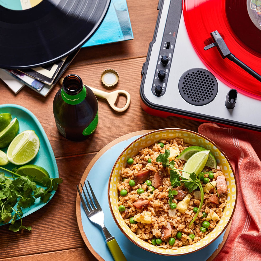 a tabletop view of a delicious bowl of BBQ pork fried rice sitting amidst records and a record player with a bright red vinyl on the turntable. There is an open beer and a plate of limes and cilantro next to it.