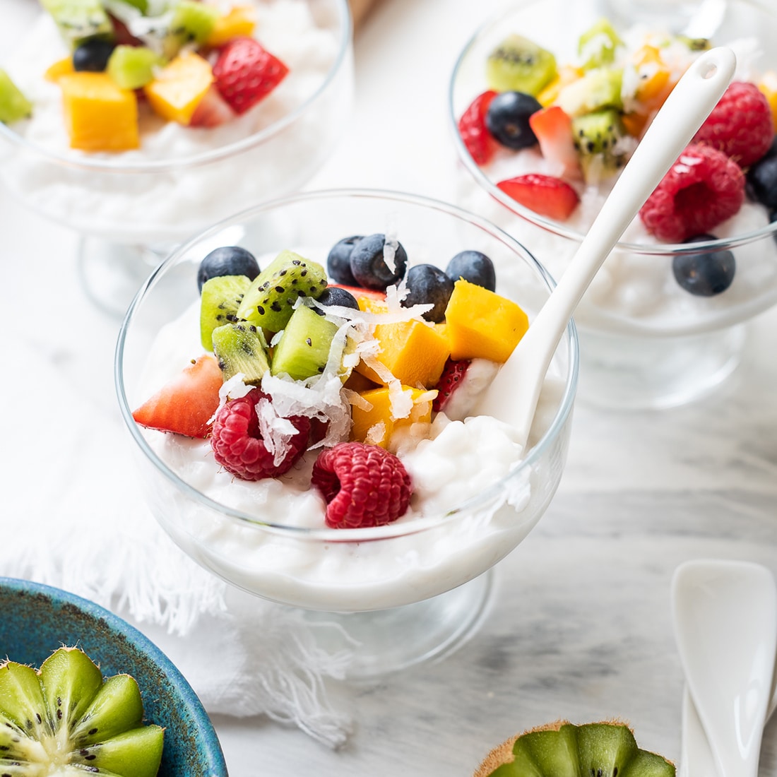 A close up of coconut rice pudding in a small delicate desert glass. There pudding is topped with fresh fruit and berries and some shaved coconut. There are two more dishes in the background, for you and friends to enjoy!