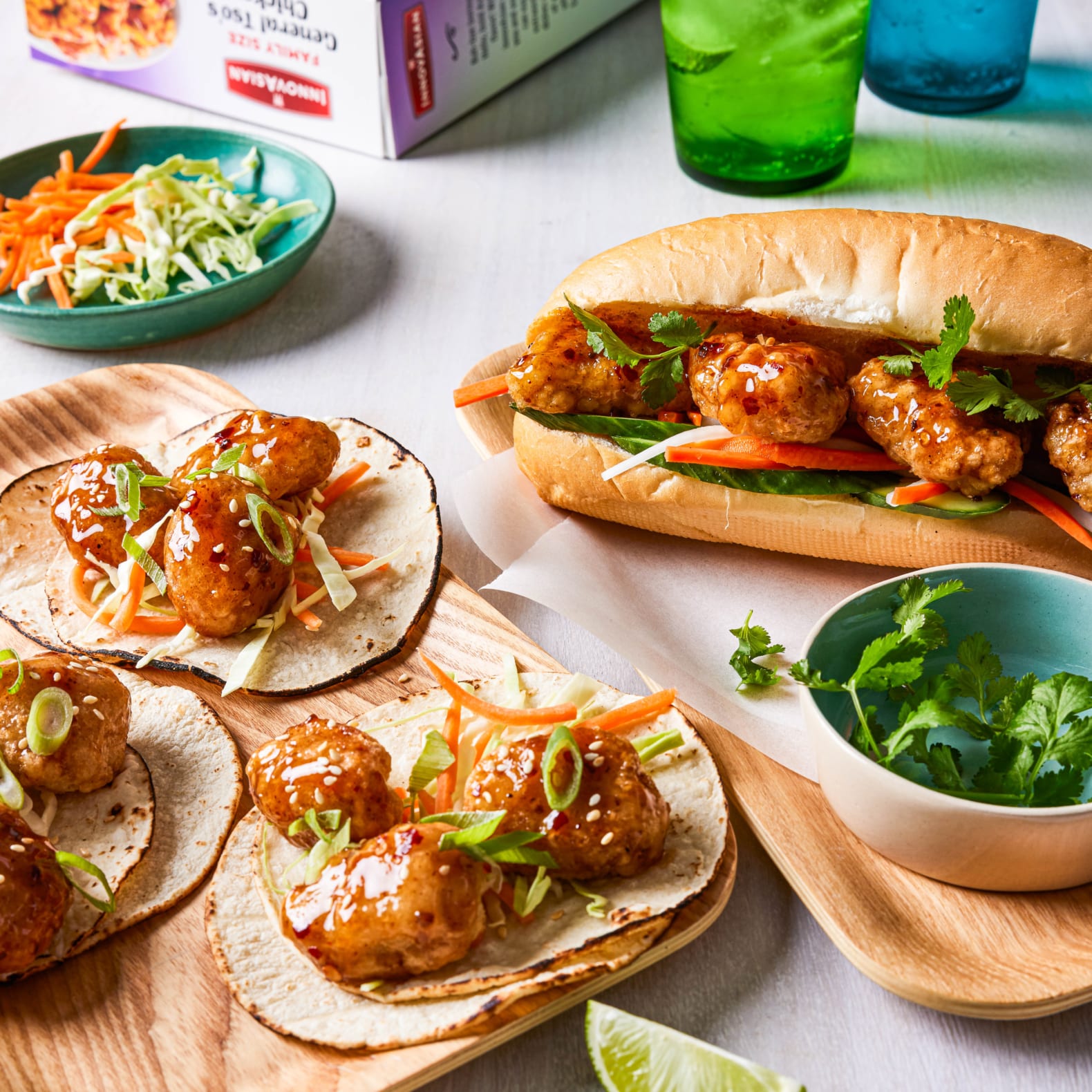 General Tso's Chicken prepared in two different ways: one as a Bahn Mi sandwhich and the other as small street tacos. Around them are cut vegetables and garnishes and the box of InnovAsain General Tso's Chicken, showing they were just prepared.