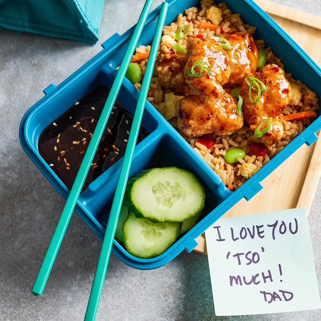 A close-up of a colorful bento box lunch containing InnovAsian General Tso's Chicken over Vegetable Fried Rice with cucumbers and dipping sauce in other sections of the box. A post-it note next to it reads "I love you 'Tso' much! - Dad"