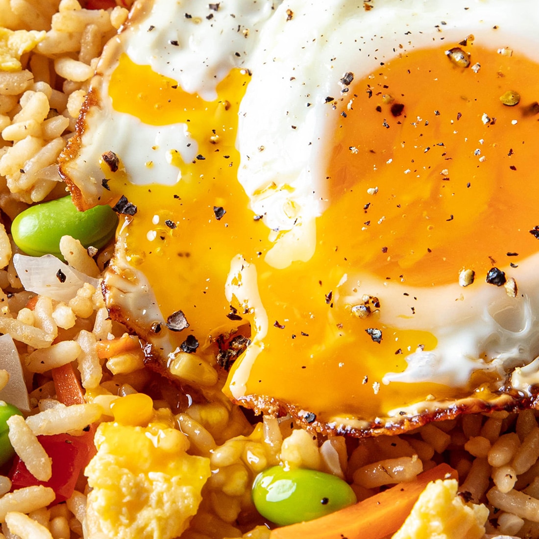 A super close-up of an amazing sunny side up egg over InnovAsian Vegetable Fried Rice. The edges are crispy and the yolk is broken and running down into the rice, seasoned with pepper and furikake.