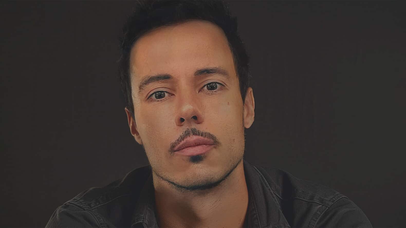 Portrait of a person with short dark hair and anchor beard mustache. They stare into the camera thoughtfully.