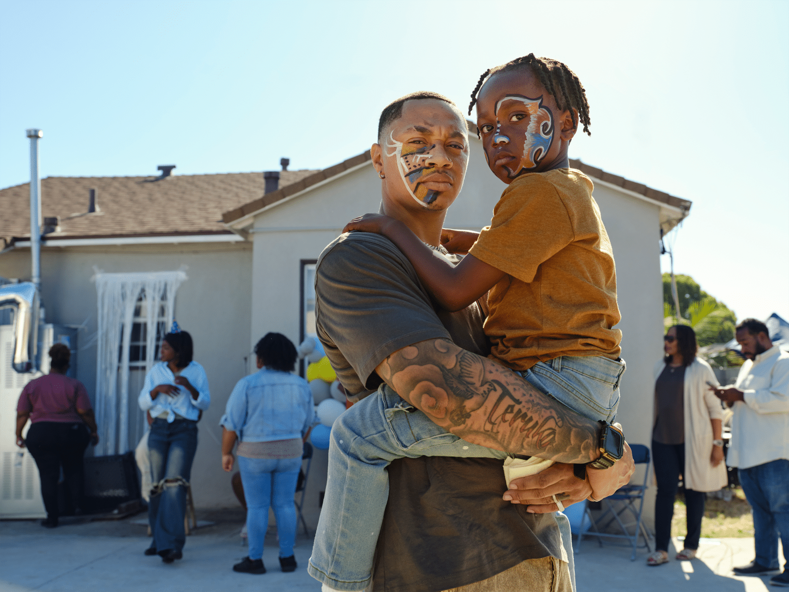 A photo of Isaiah, a father in his 20s, holding his 7-year-old son, Isaiah Jr. Both are looking down at the camera with a powerful gaze. They are in the sunny backyard of their Compton home. Friends and family are sprinkled around the backyard, with blue and yellow balloons as if they are all there to celebrate someone’s birthday.