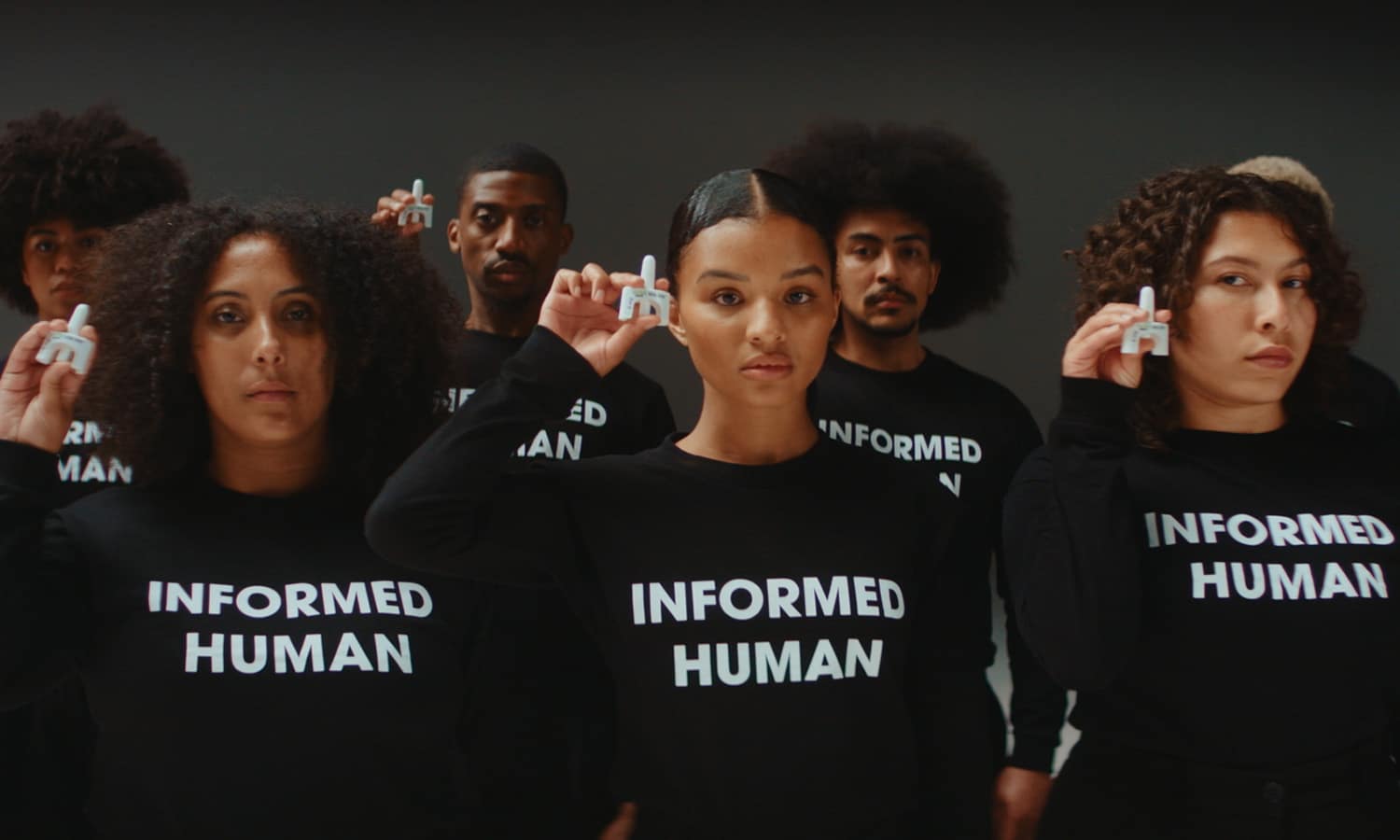 Two lines of young people in a dark, plain room looking directly to camera and holding up containers of naloxone. They are wearing matching black shirts that say “INFORMED HUMAN” across the chest. Their facial expressions are serious and powerful.