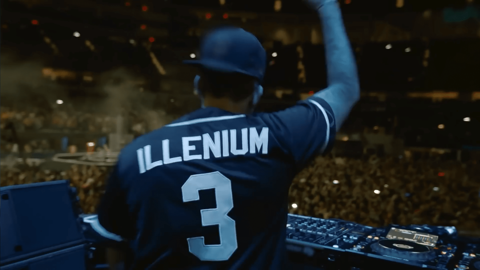 EDM artist Illenium as seen from behind playing a show in a large concert venue. He is wearing a sports jersey style shirt with “ILLENIUM” and the number 3 on his back. He is raising his arm to the crowd in an energetic way.