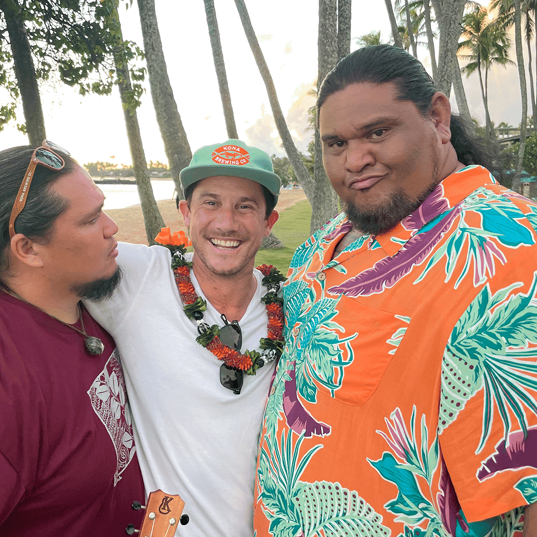 Three men pose under palm trees, Andy smiling in a hat and lei, flanked by Brutus and David.