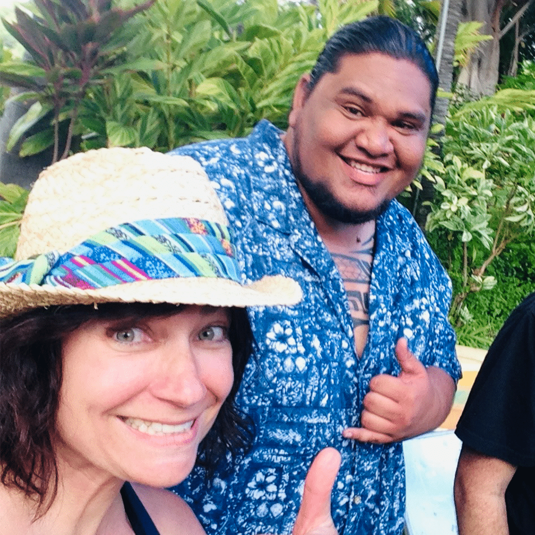 Anne in a straw hat smiles at the camera, with David in a blue Hawaiian shirt giving a shaka sign behind her.