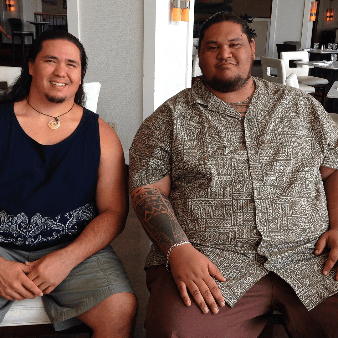 Two men sit side-by-side, Brutus in a dark tank top and David in a patterned short-sleeved shirt with a tattooed arm.