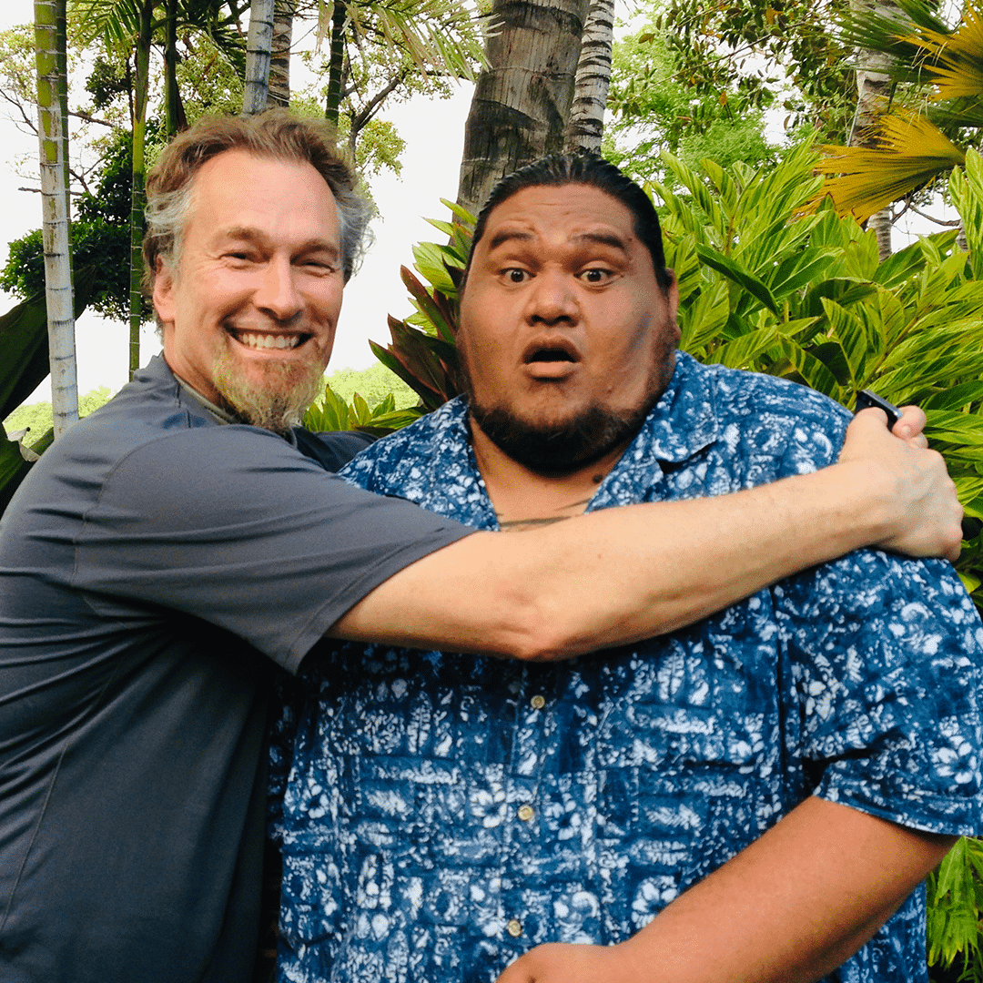 A man with a beard and gray shirt hugs David in a blue Hawaiian shirt, who looks surprised.