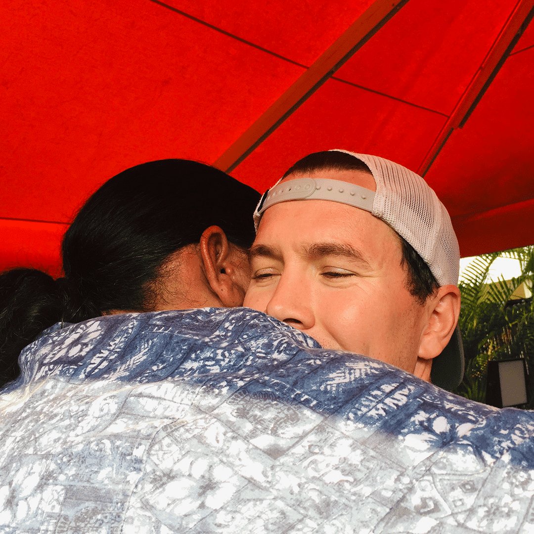 Keenan, in a baseball cap with closed eyes, hugs David whose back is to the camera, under a red umbrella.